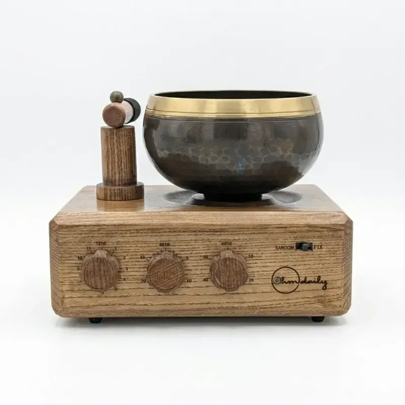 The second-generation unit from the front: a dark hand-hammered brass singing bowl with a gold rim on an ash hardwood base, a wooden mallet on a post at the left, and three wooden dials with a Random/Fix switch on the front panel